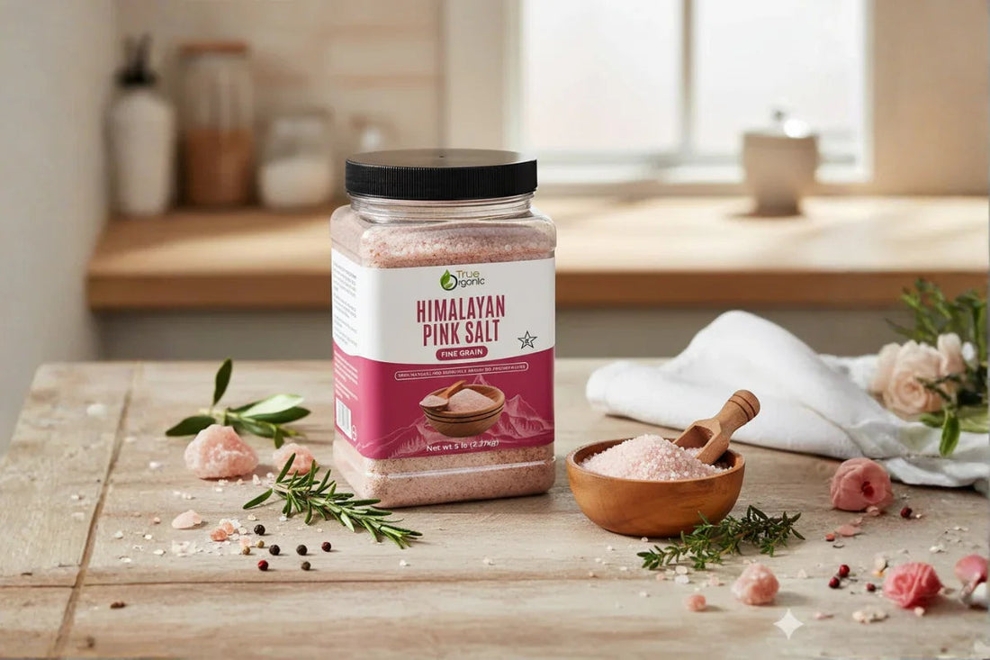 Container of True Organic Himalayan pink salt with wooden bowl and herbs on kitchen table